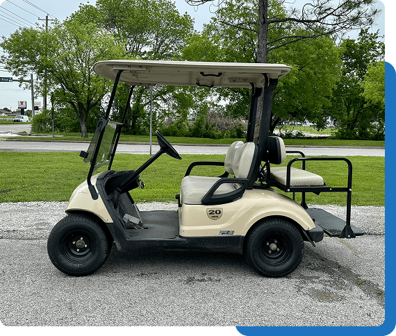 A beige golf cart parked on gravel near trees and grass.